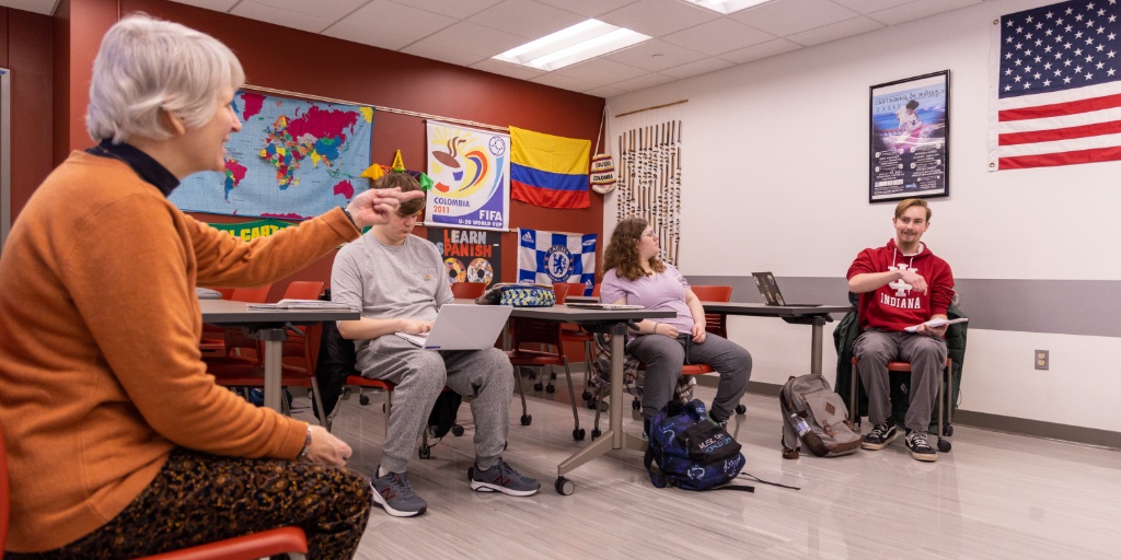 An instructor in an orange sweater points at three students.